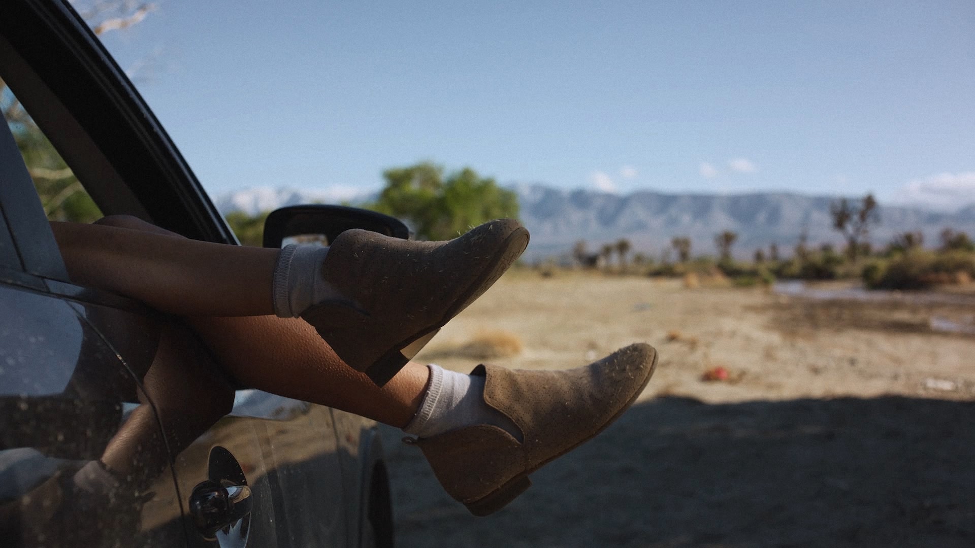 Une personne assise dans une voiture garée, les pieds chaussés de bottes dépassant par la fenêtre, avec des montagnes en arrière-plan.