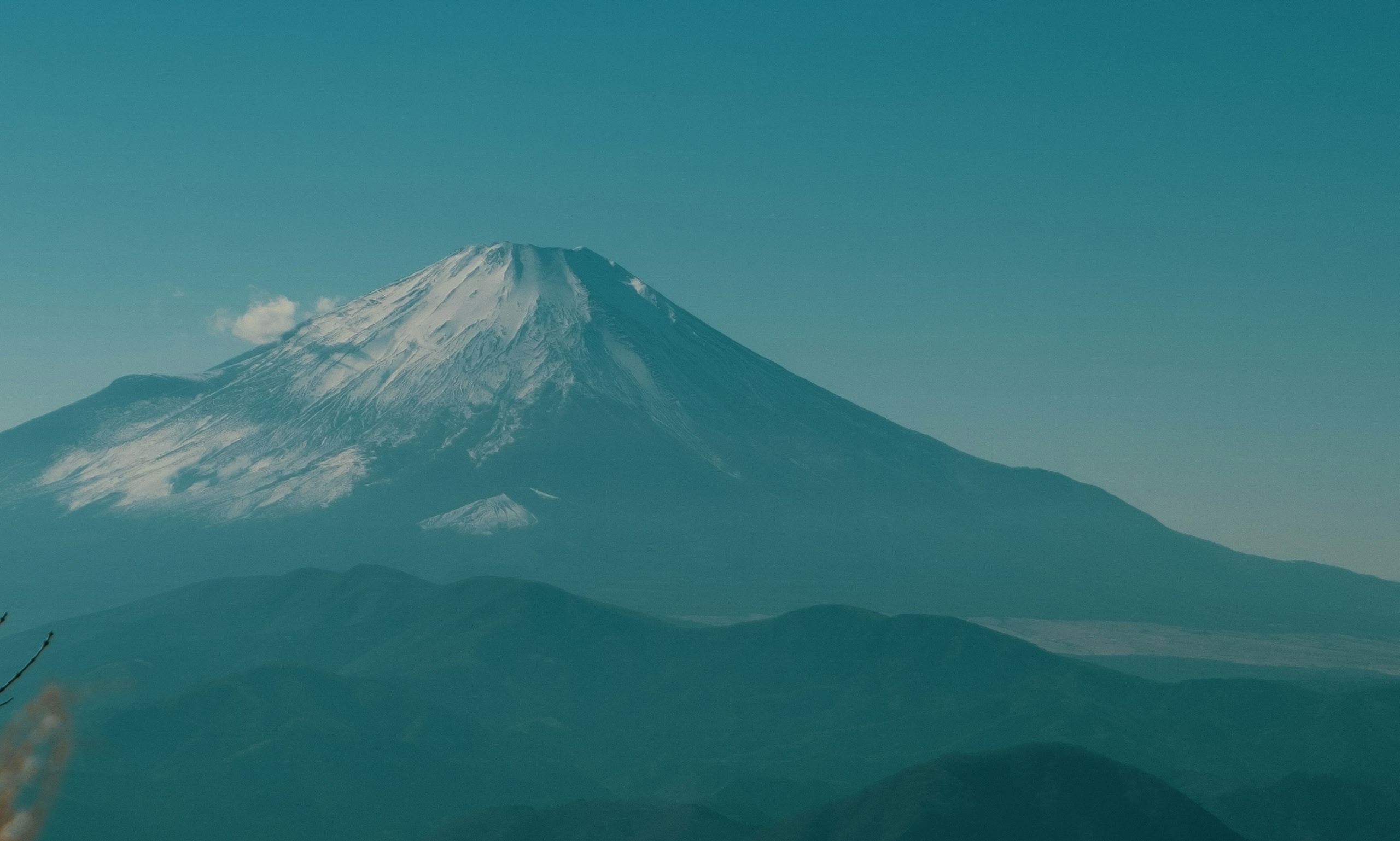 Snow-capped Mount Fuji rises above hazy mountain ridges beneath a blue sky.