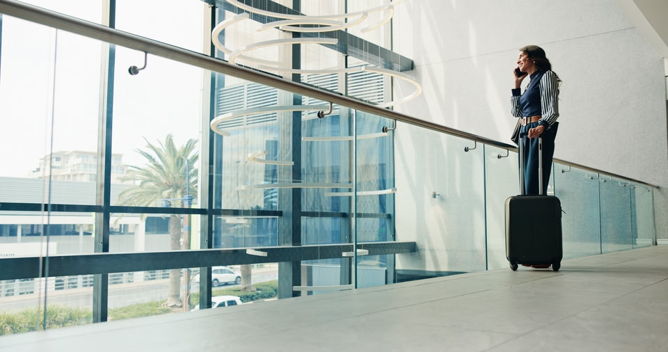 A professional traveler stands next to a rolling suitcase on a mezzanine overlooking a large window while talking on the phone.
