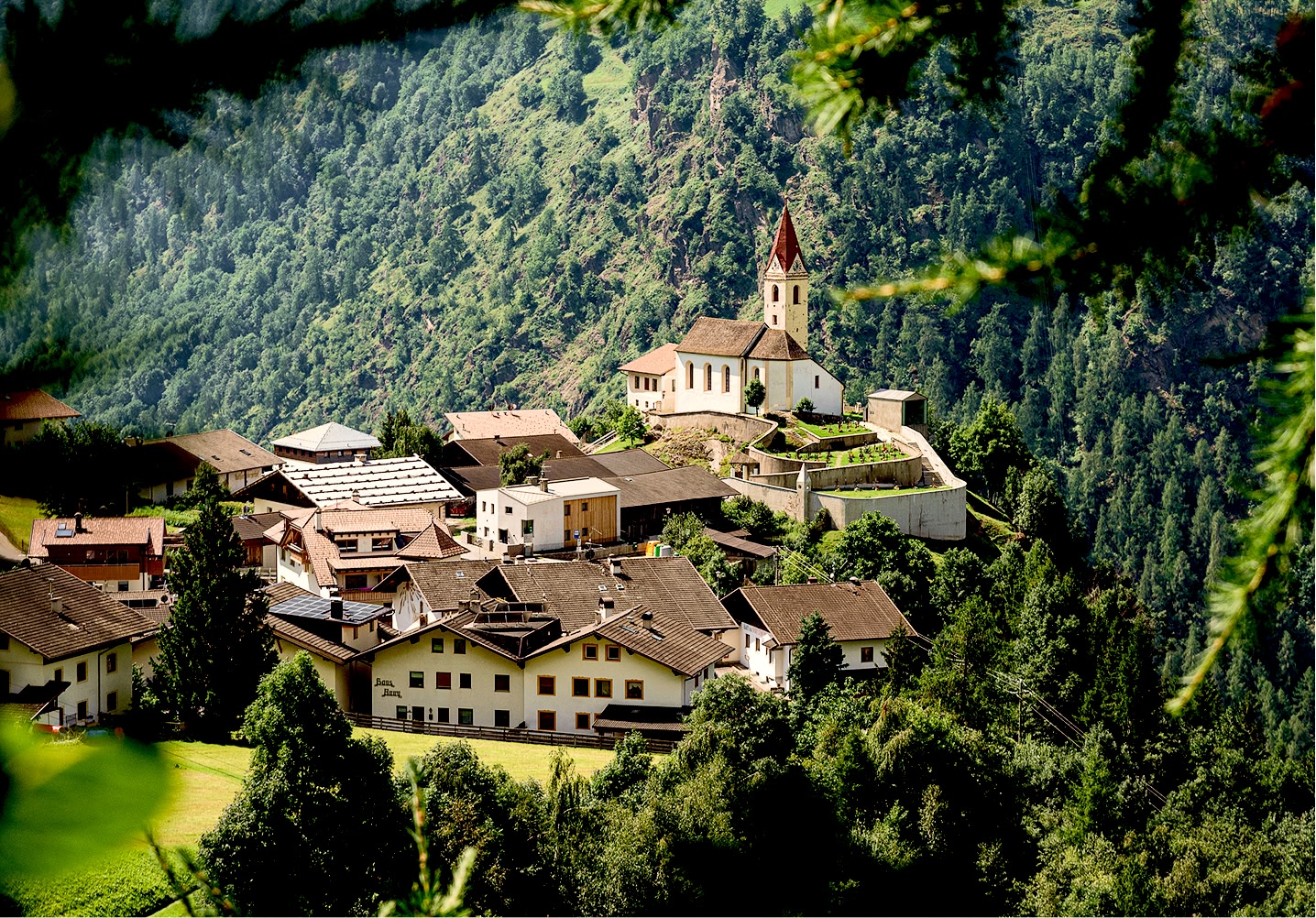 Alpendorf mit einer Kirche auf einem Hügel, umgeben von bewaldeten Bergen.