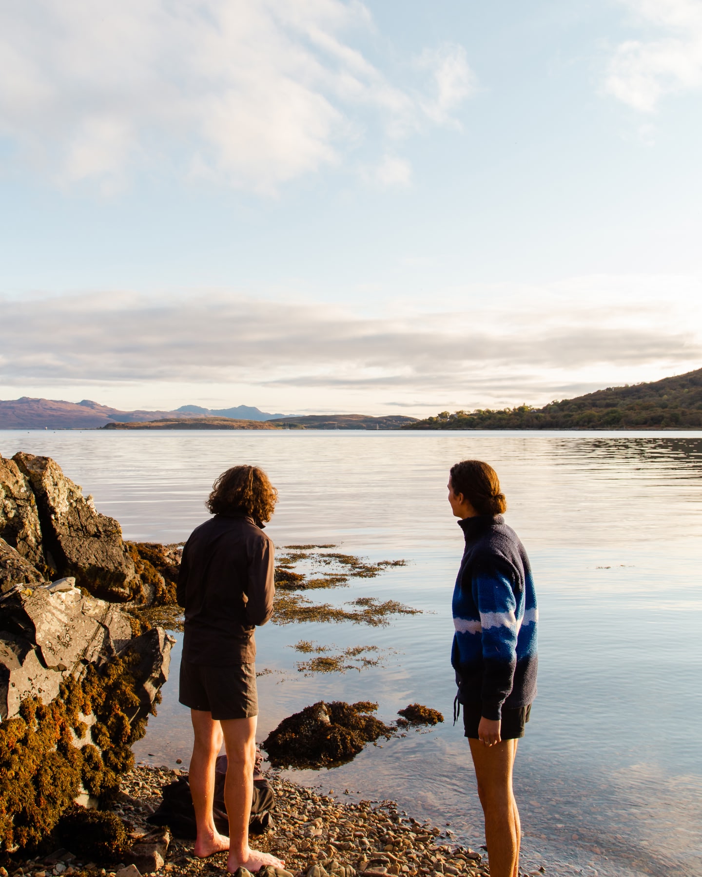 Two people standing on a rocky shoreline overlooking a calm bay.