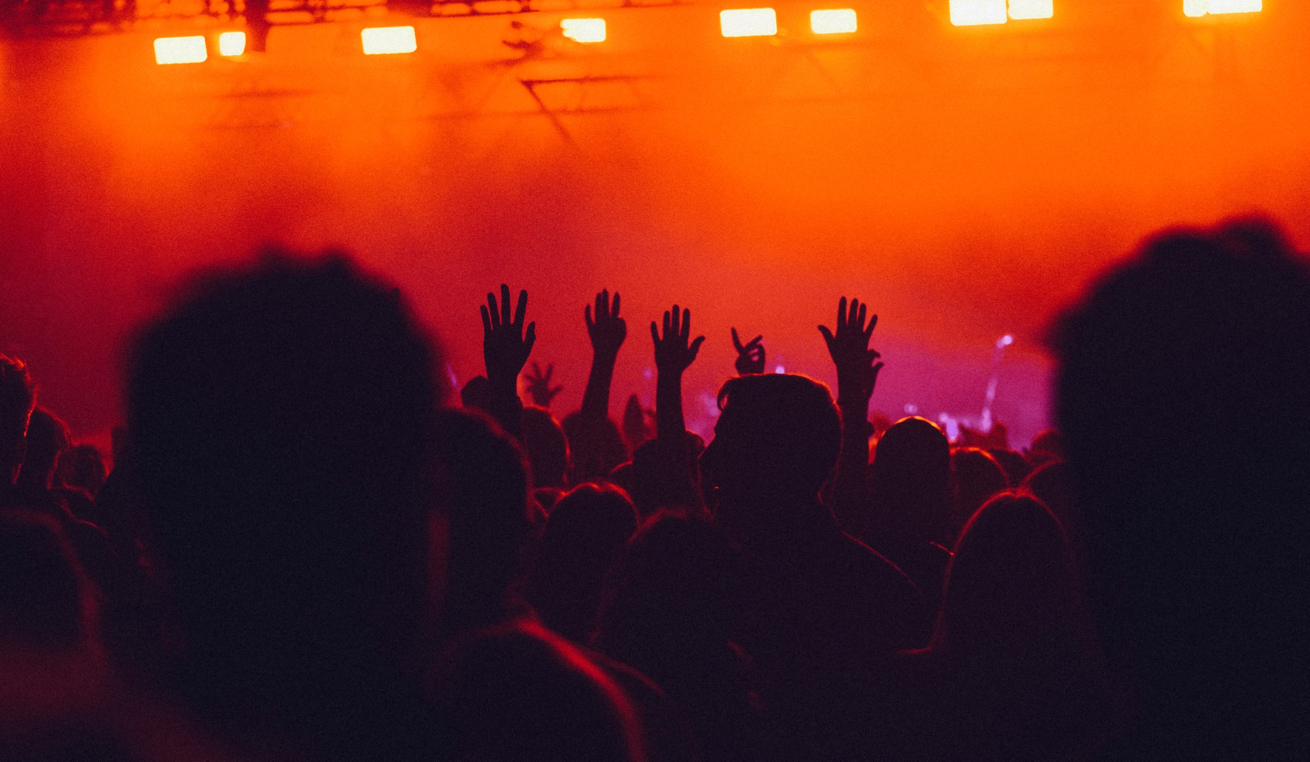 Silhouettes d’une foule de festival, les mains levées, sur fond de scène aux couleurs orange, rouge et violette éclatantes.