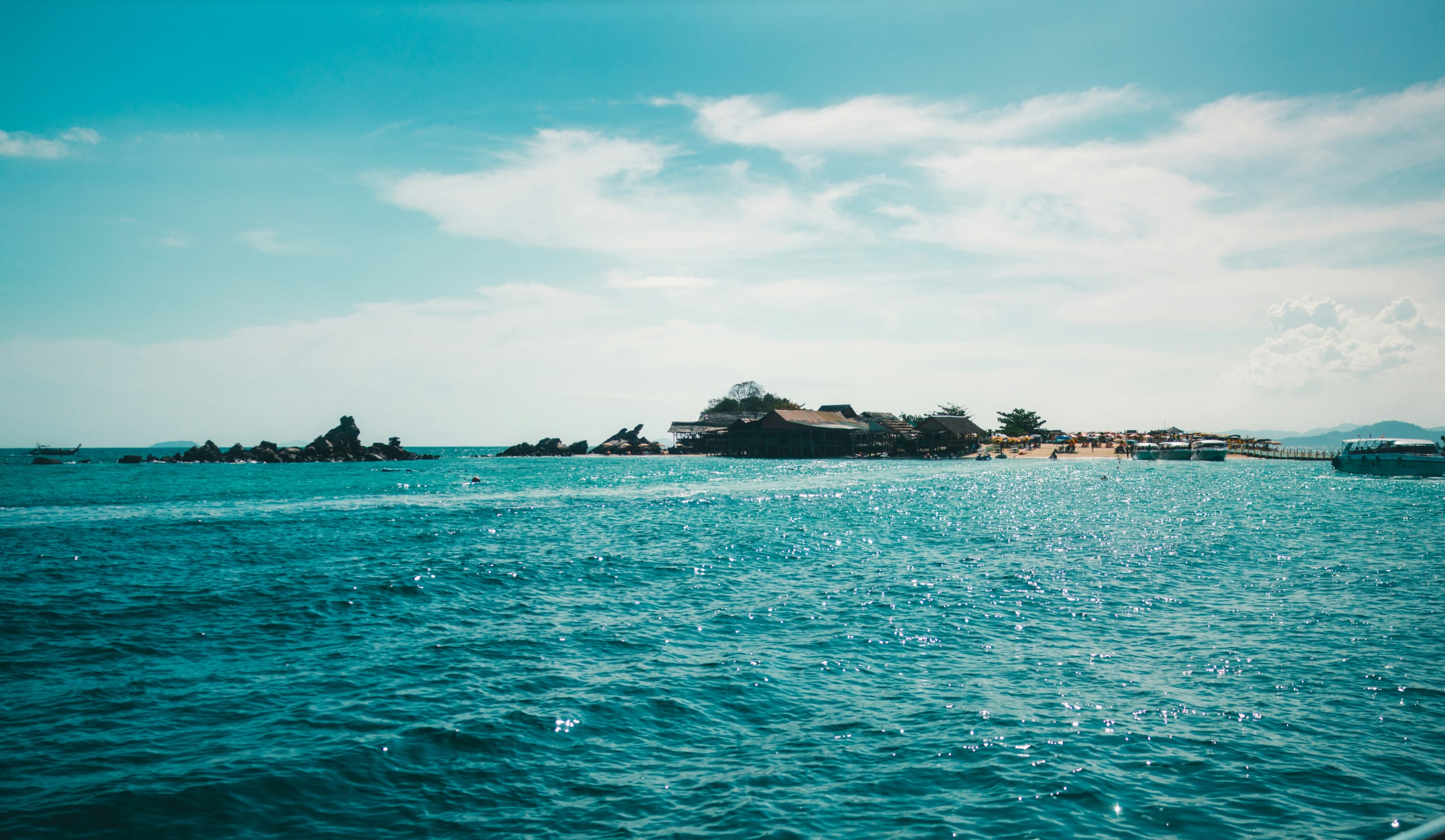 Turquoise sea with rocky outcrops and a tropical island resort featuring overwater buildings, a sandy beach, boats, and a bright blue sky.