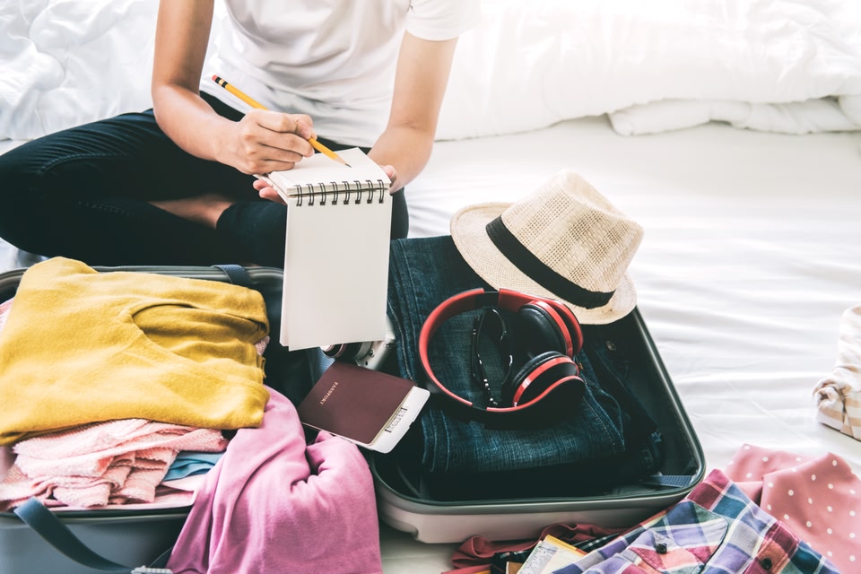A person sits on a bed near an open suitcase while writing a checklist in a notebook.   