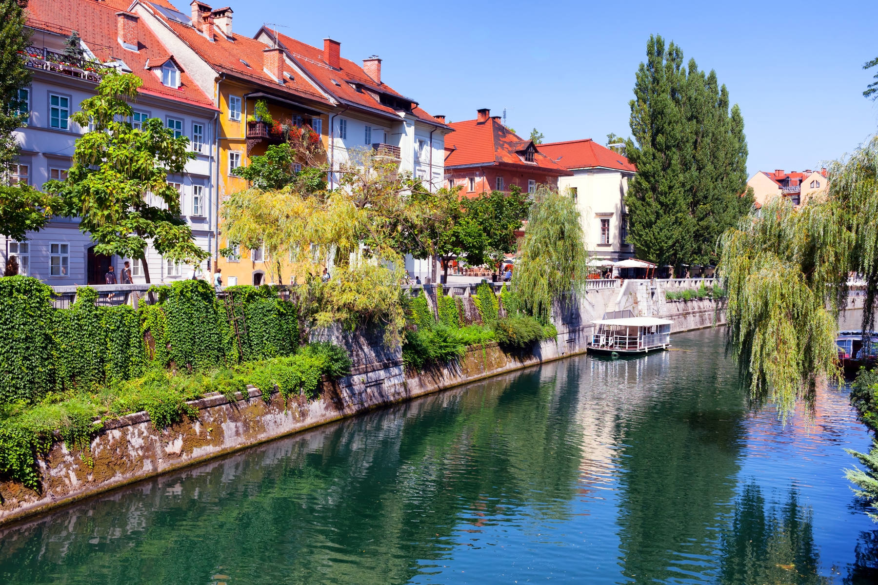 Photo shows traditional houses in Ljubljana, the capital of Slovenia, one of our unexpected destinations. The sun is shining, the houses are next to a river or canal with a boat on it, lined by trees.