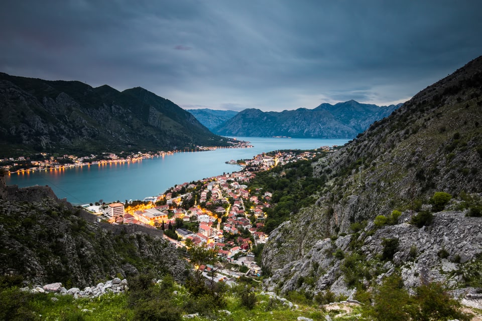 Kotor by night. Tree-covered mountains soar over a winding lake, while the shimmering town is nestled at the base.