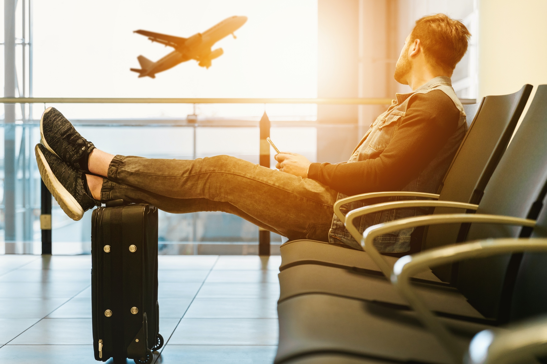 A man sits with his feet on his suitcase while a plane takes off in the background.