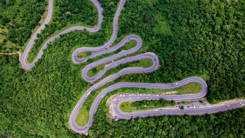 Aerial view of a winding mountain road with multiple sharp hairpin turns cutting through dense green forest.