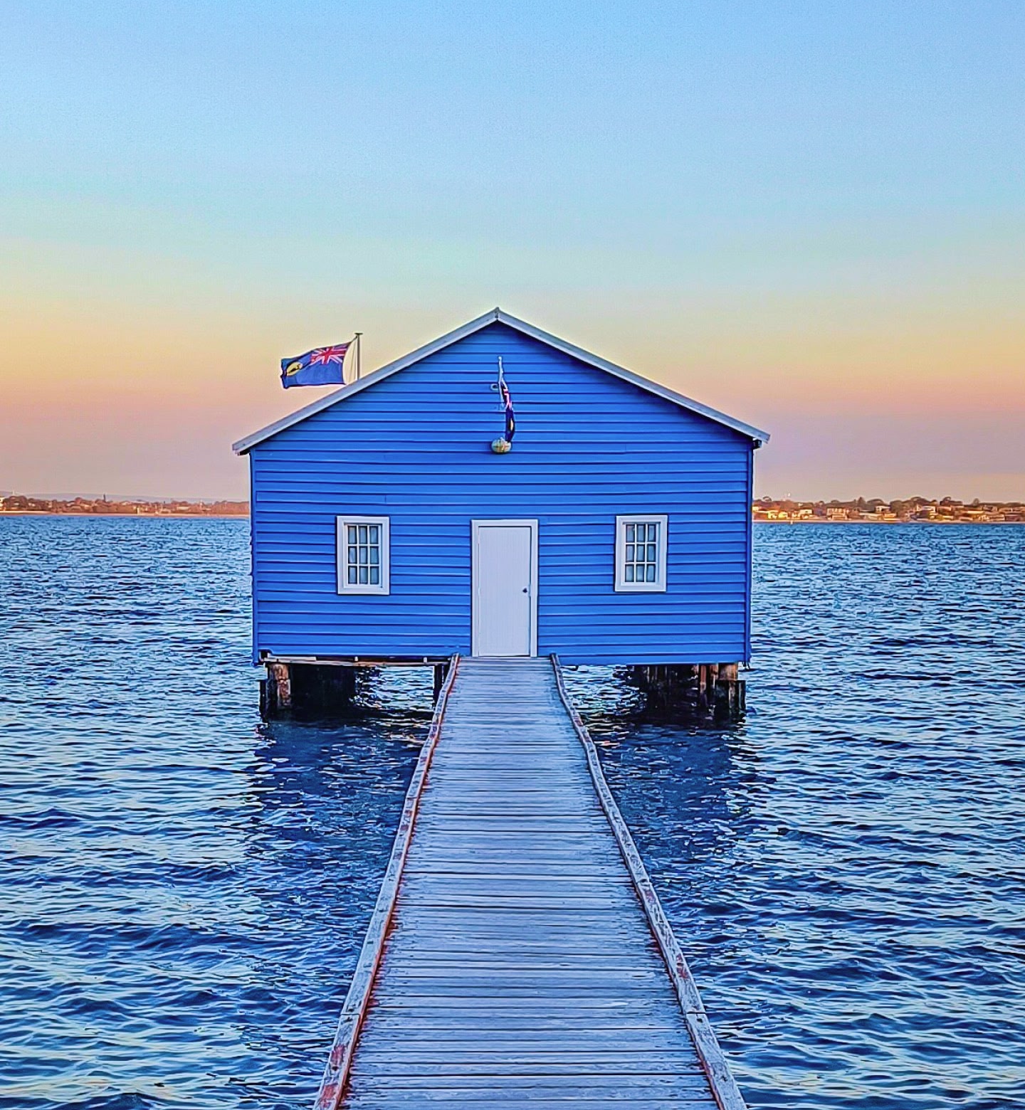 A wooden jetty leads to a bright blue boathouse on the water in Perth, Australia.
