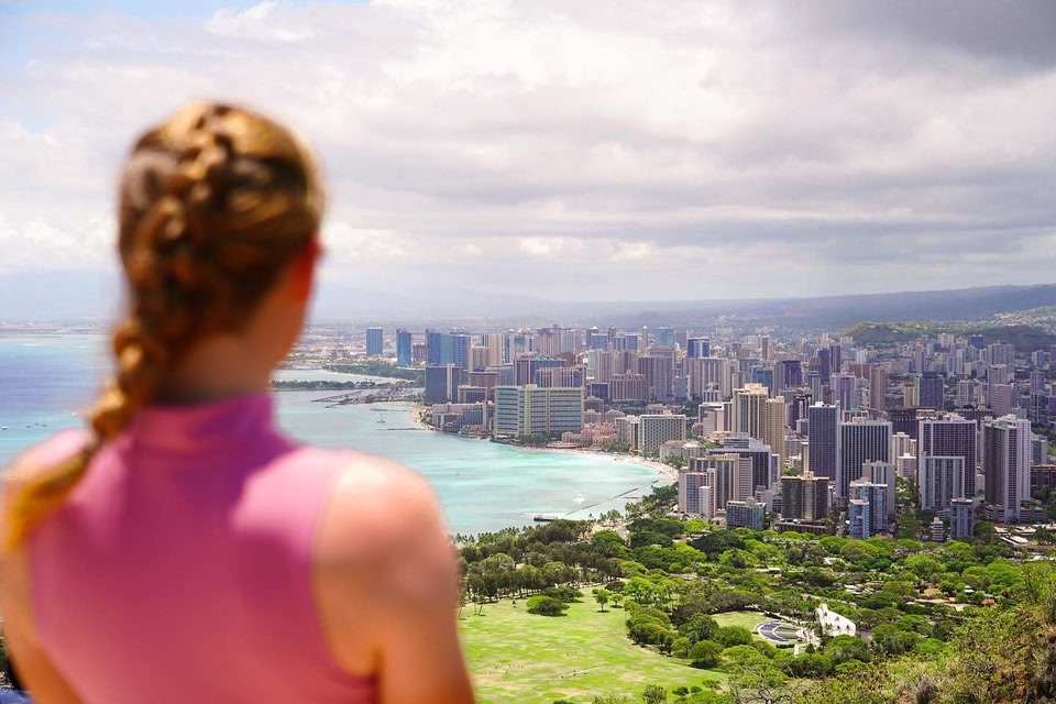 A woman shown from behind with braided hair is looking out at a panoramic view of a coastal city skyline bordering a turquoise bay under a cloudy sky.