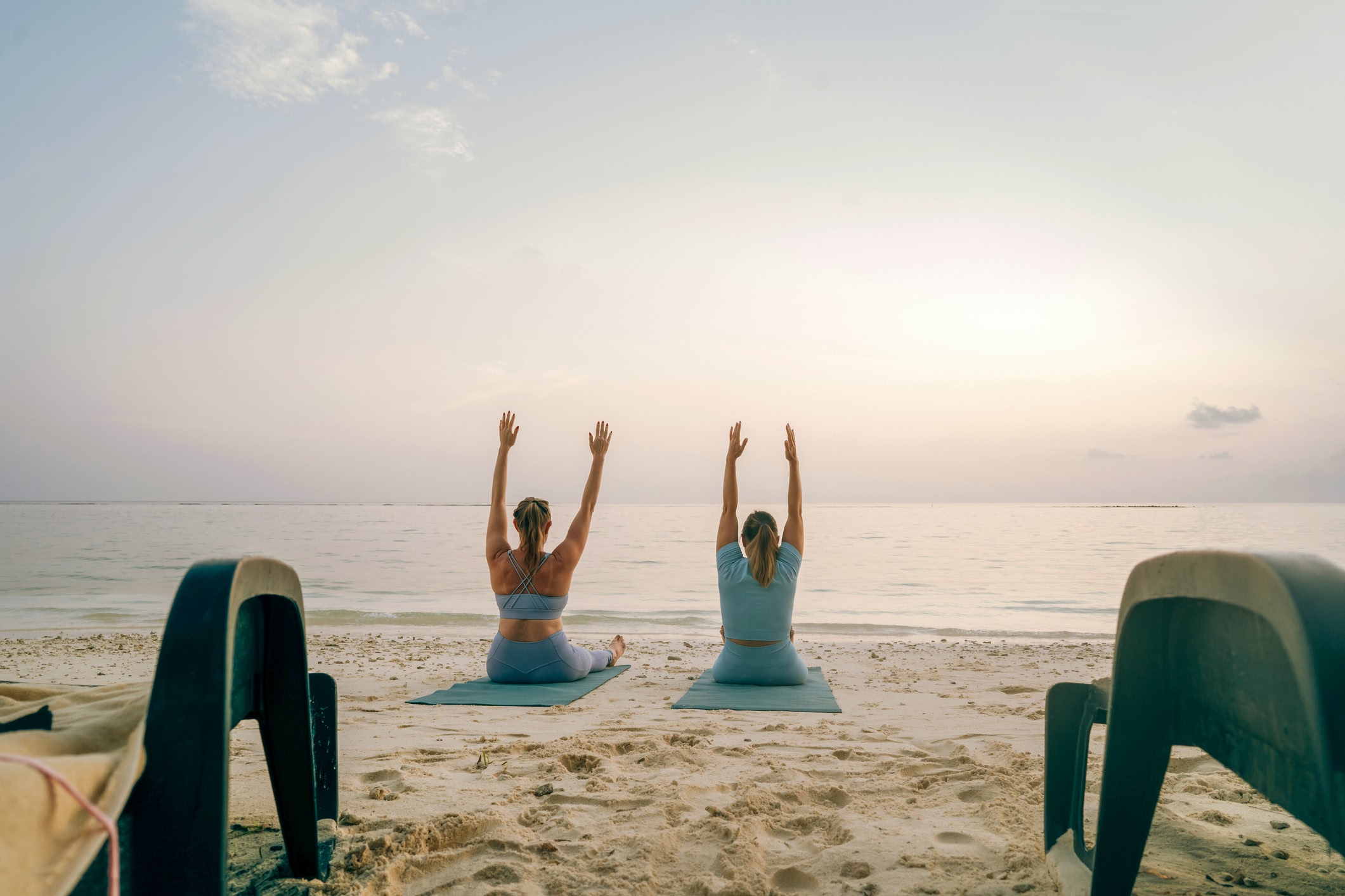 Een brede opname van twee vriendinnen die op een tropisch strand zitten met uitgestrekte armen en uitkijken over de Indische Oceaan, uitzicht vanaf de achterkant. Ontspanning, mindfulness en zen-achtig, yogales, pilates, stretchen, energiebeoefening.