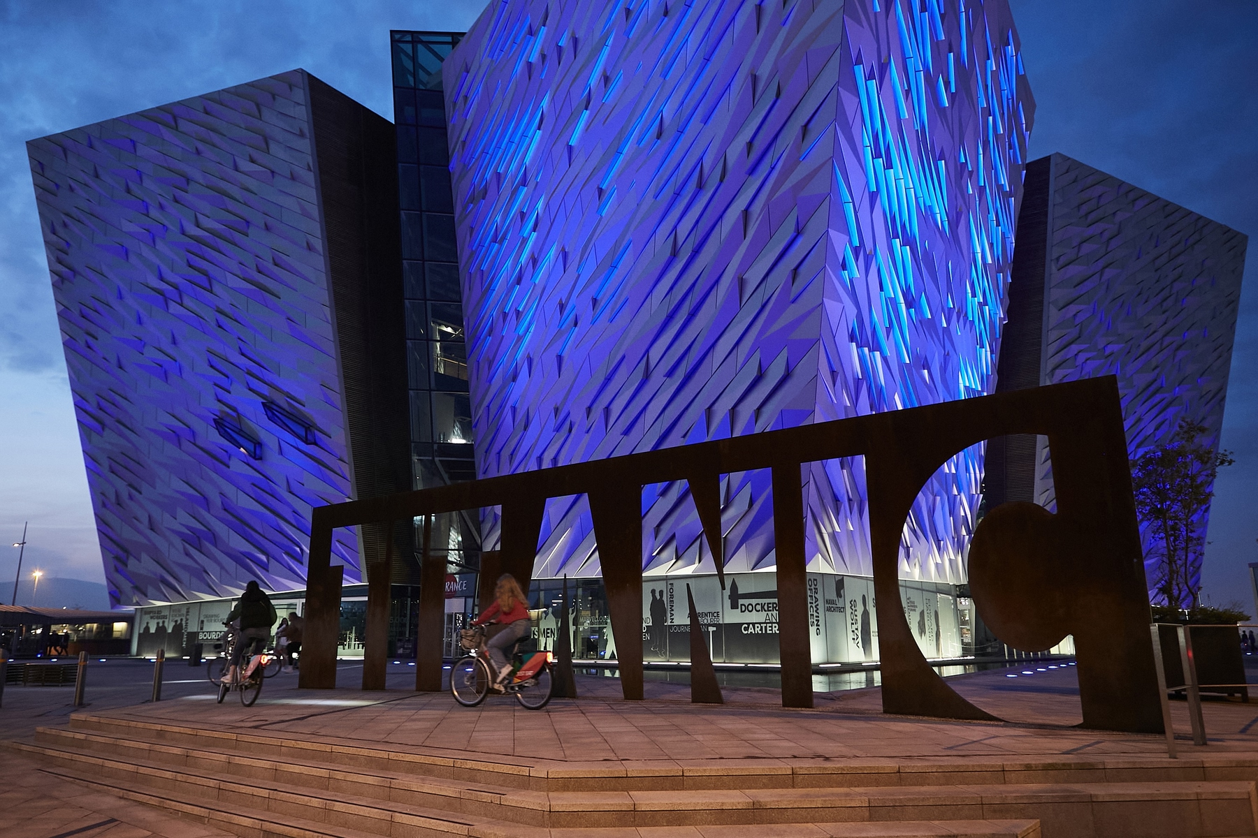 The Titanic Belfast museum building at night, with two people cycling past.