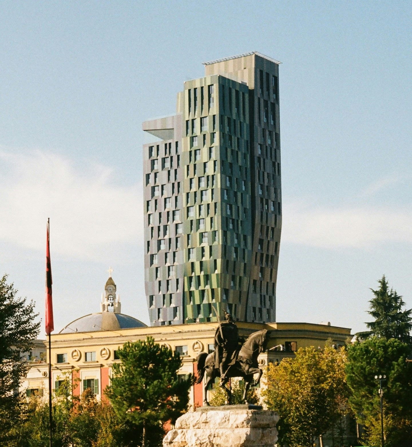 Uma torre verde moderna atrás da estátua de um cavaleiro e da bandeira albanesa em Tirana, Albânia.