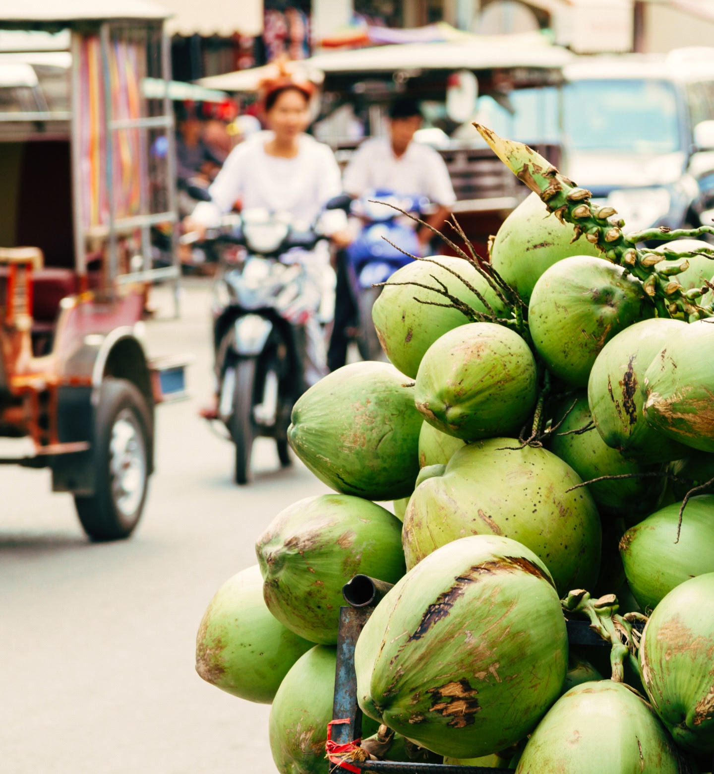 Two people riding mopeds in busy traffic with green coconuts in the foreground in Phnom Penh, Cambodia