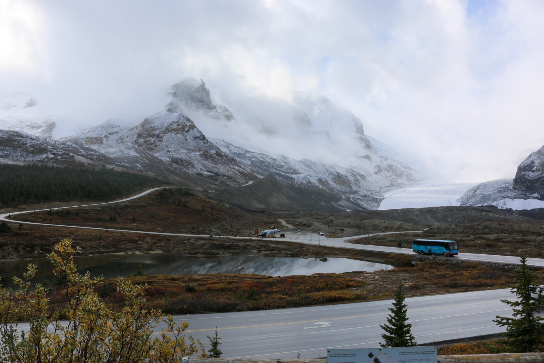 a blue bus driving along the highway with a mountain range in the background on the icefields parkway, a popular road trip in western canada