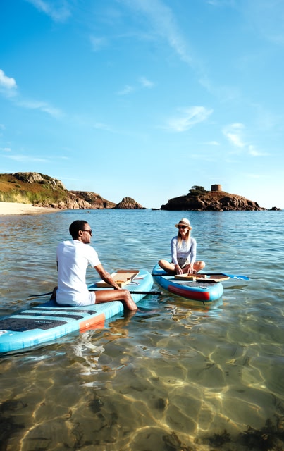 Couple sitting on paddleboards in the tranquil sea, with the white sand Jersey beaches in the background.