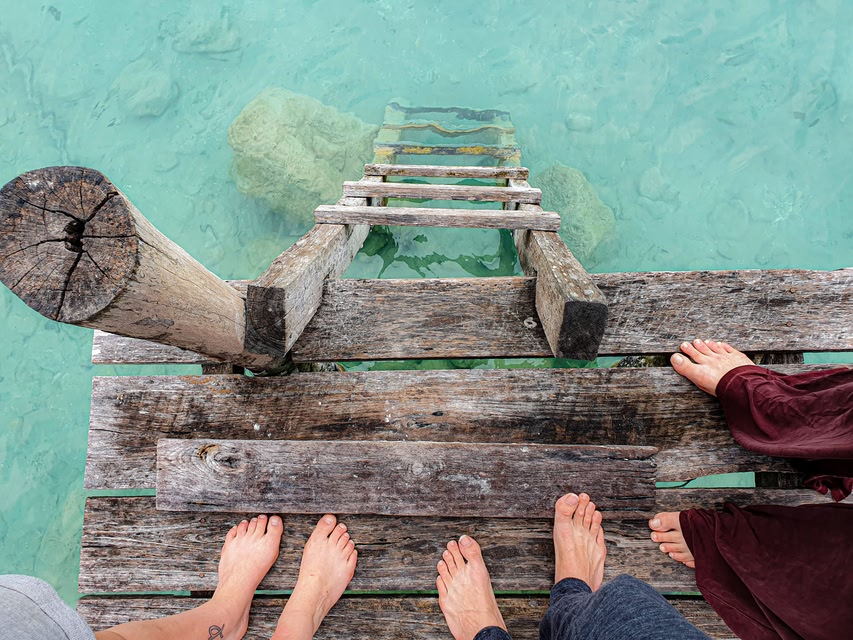 Looking down, view of people's feet standing on a wooden dock with a ladder down to clear water below.