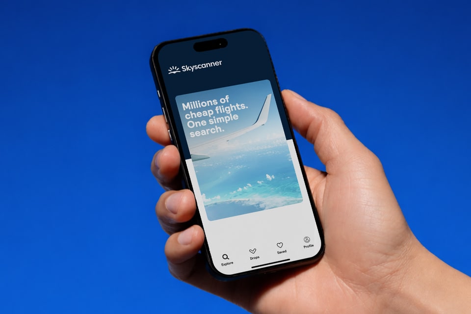 A hand holds a smartphone displaying the Skyscanner app against a solid blue background.