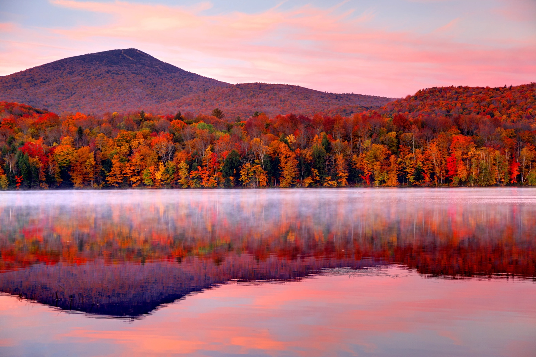 A pink sky over red autumn trees reflecting in a lake.