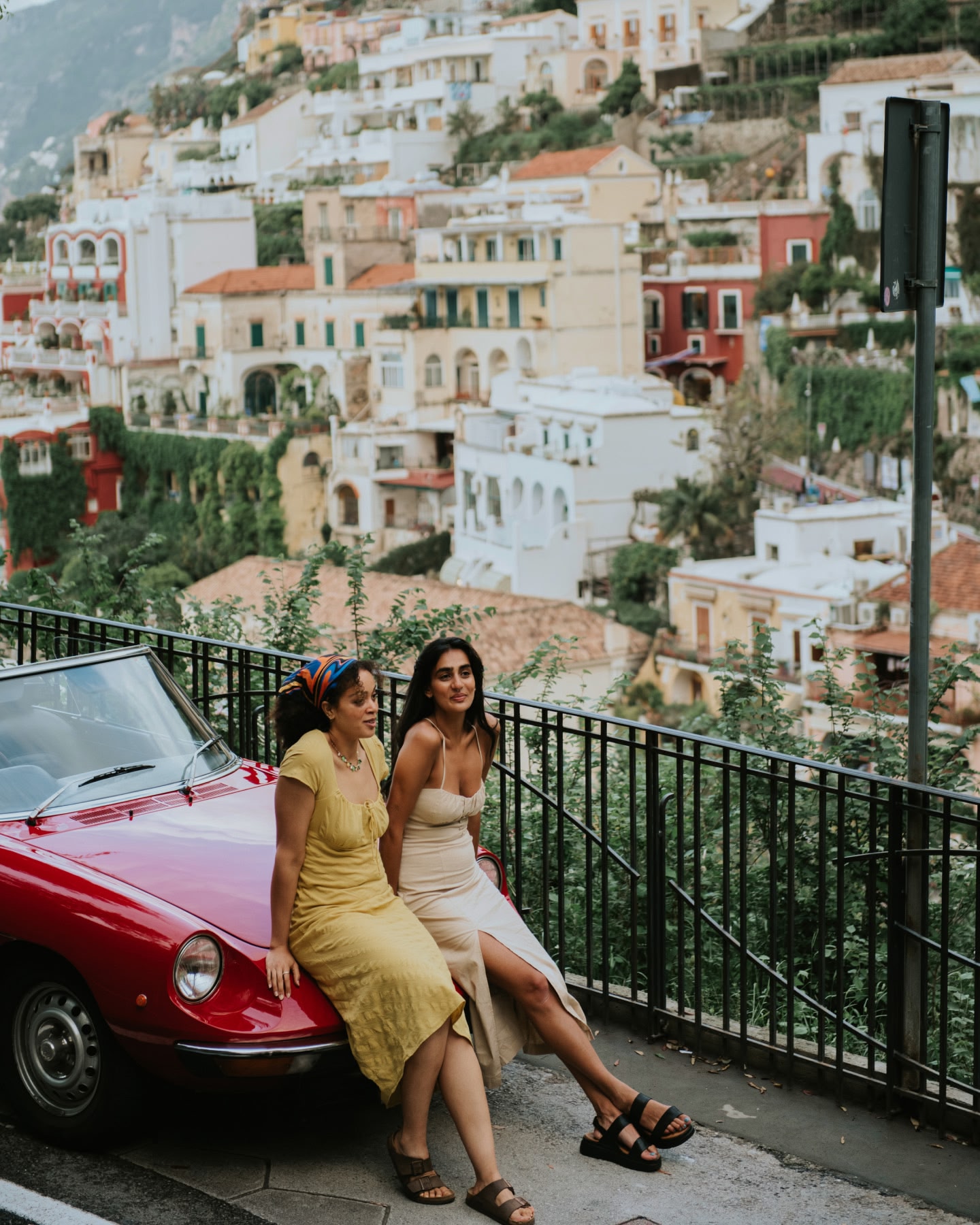 Two women sit on the hood of a red car with hillside buildings in the background.