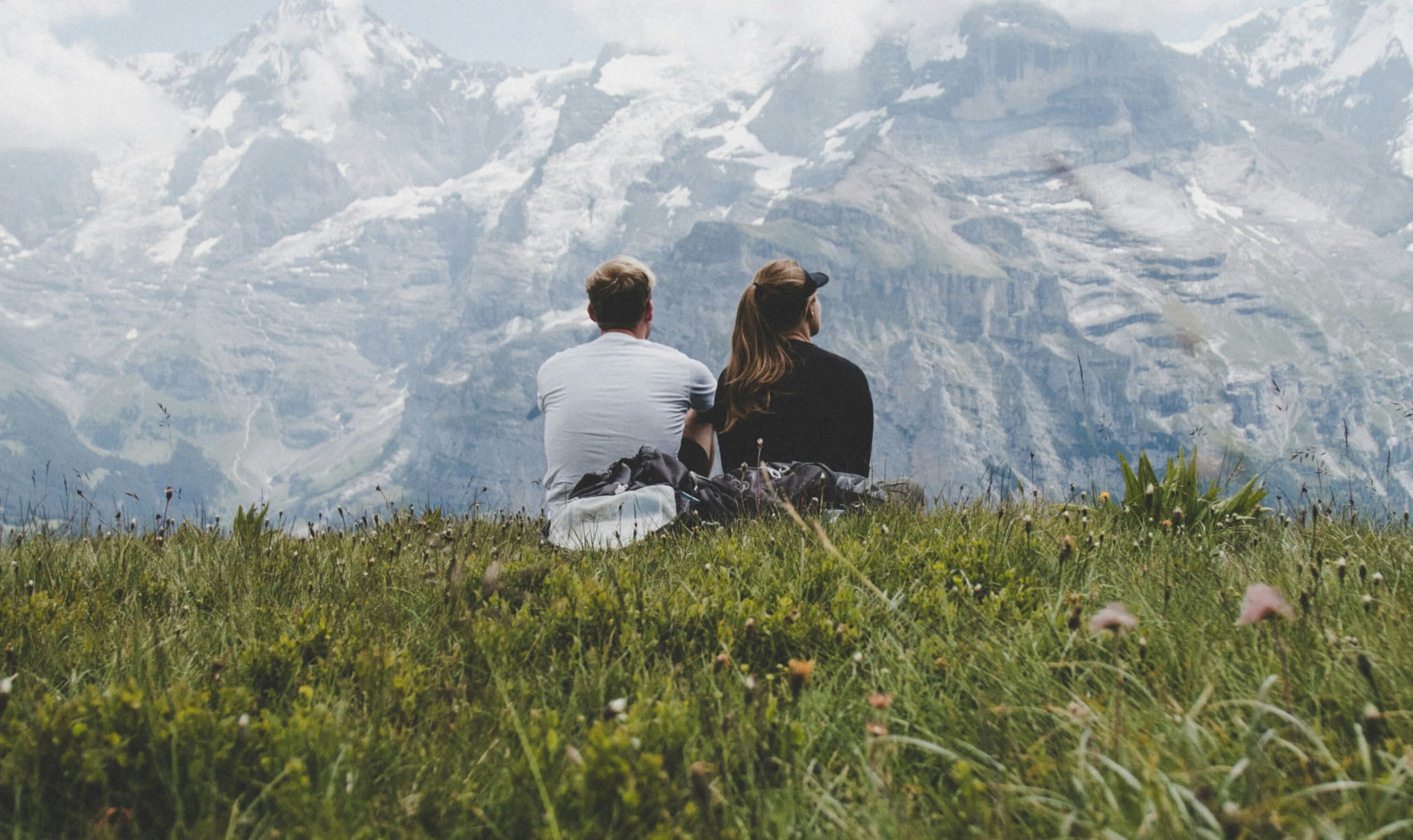 Mężczyzna i kobieta siedzący obok siebie w trawie, plecami do obiektywu, i podziwiający górski krajobraz