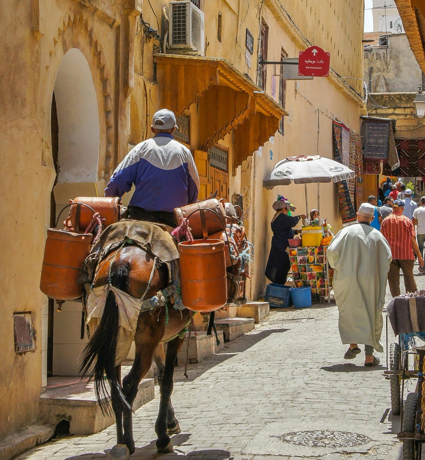 Ein Mann reitet mit mehreren Kanistern auf einem Pferd durch eine sonnendurchflutete Marktstraße in Fès, Marokko.