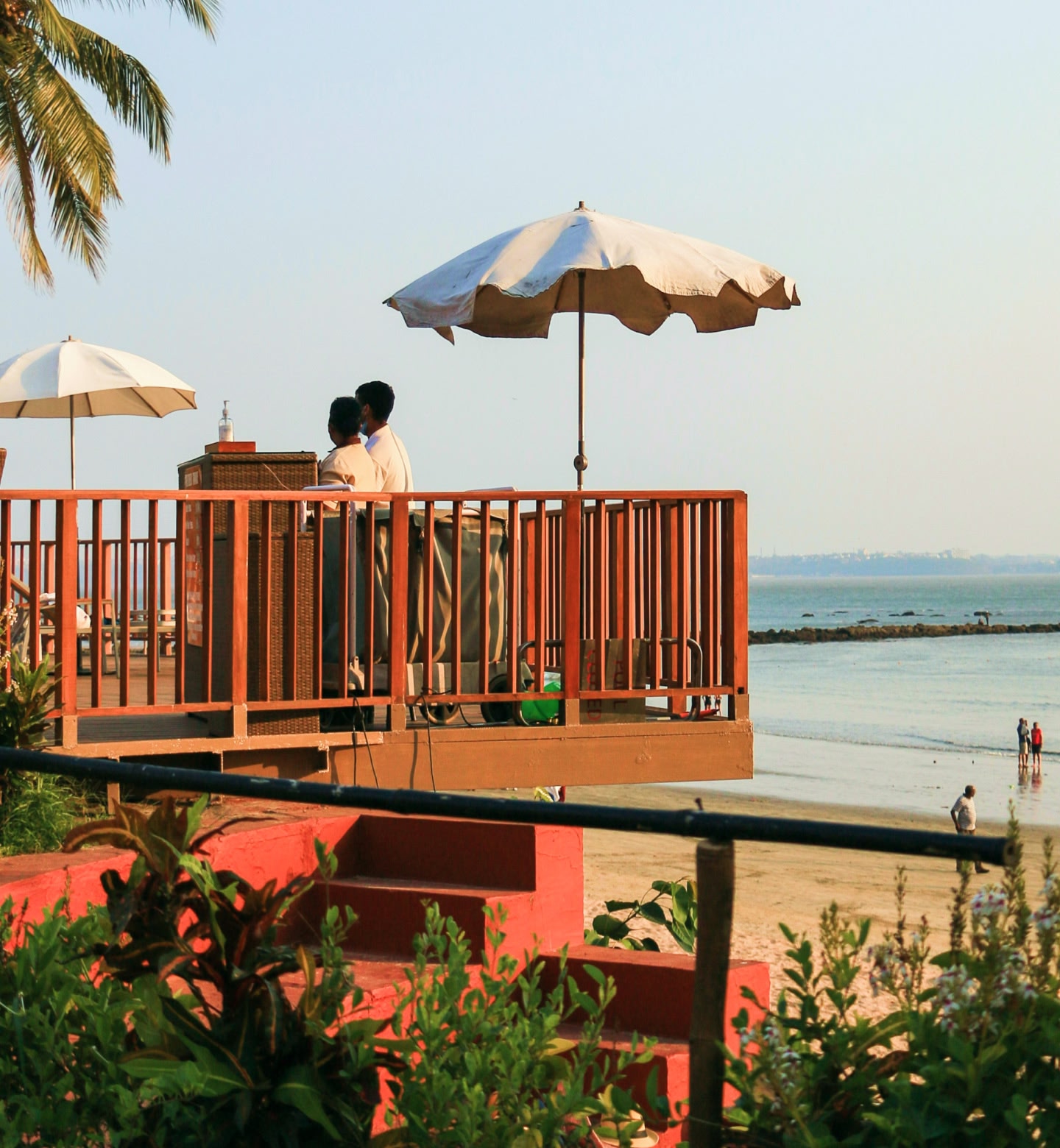 Two people standing under an umbrella on a balcony with a red railing, overlooking a beach in Goa, India