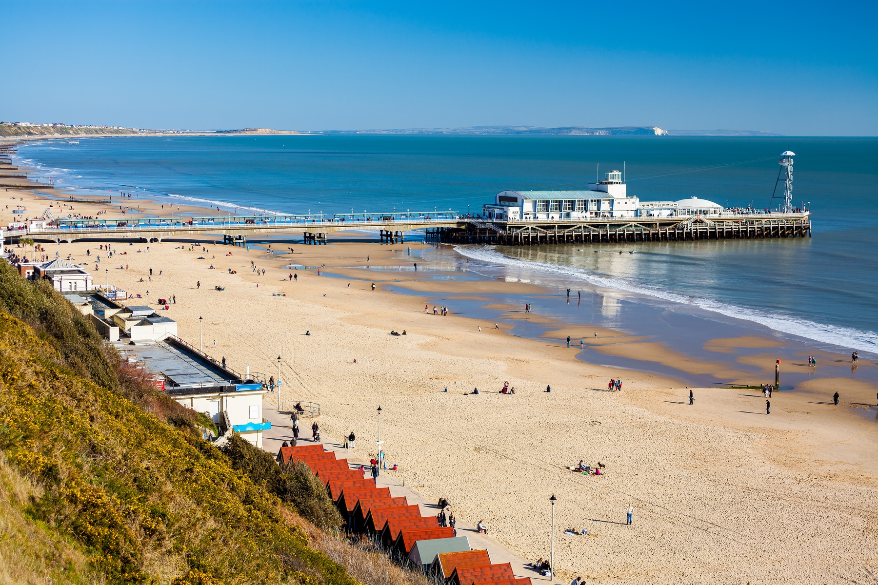 Picture shows Bournemouth Pier, the beach, and beach huts on a sunny day, with people having fun on the sand.