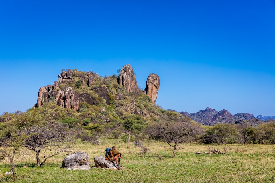 رجل يجلس على صخرة في منطقة عشبية وخلفه منظر طبيعي لتكوينات صخرية