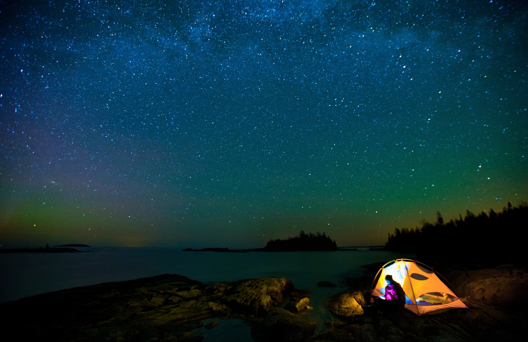 a tent lit up at night with a camper inside sitting by the rocky coastline in Atlantic Canada, under a starry night sky