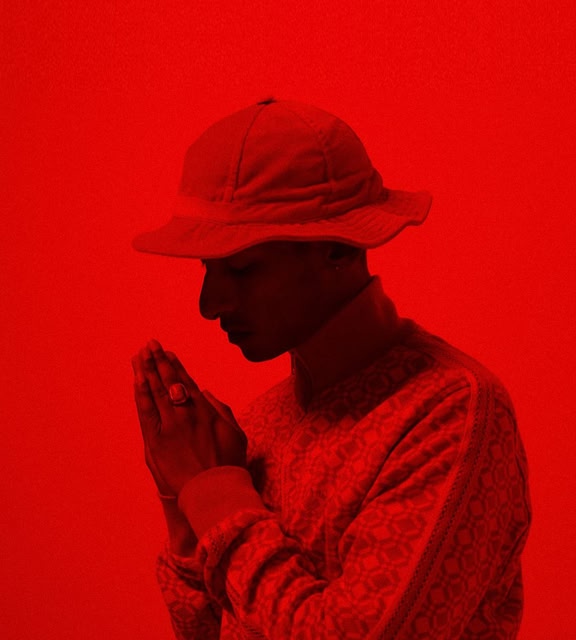 Singer Jitwam wearing a hat with his hands in the praying position. A bright red filter is over the image