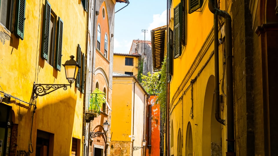 Una stretta via di una città italiana con edifici dai colori vivaci giallo e arancione, persiane verdi e una lanterna decorativa montata sulla parete