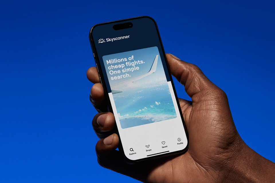 A hand holds a smartphone displaying the Skyscanner app against a solid blue background.