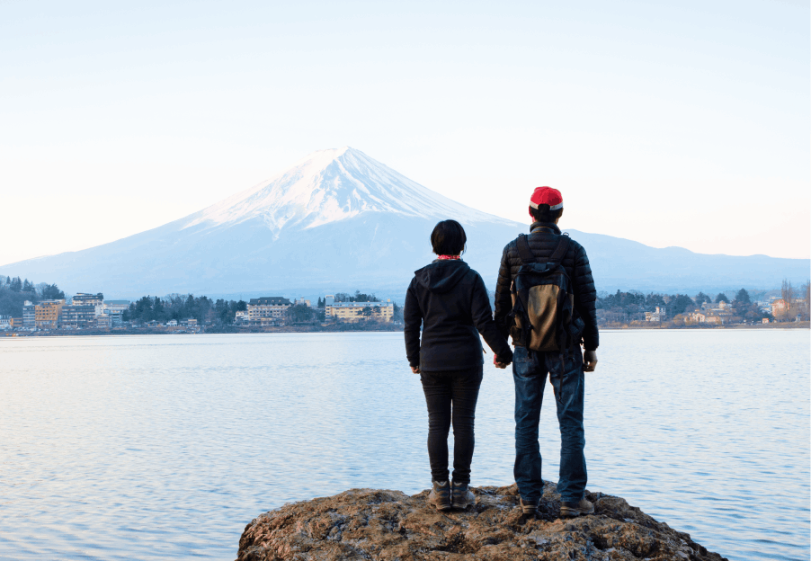 A couple holding hands and standing on the edge of a body of water looking at Mount Fuji.
