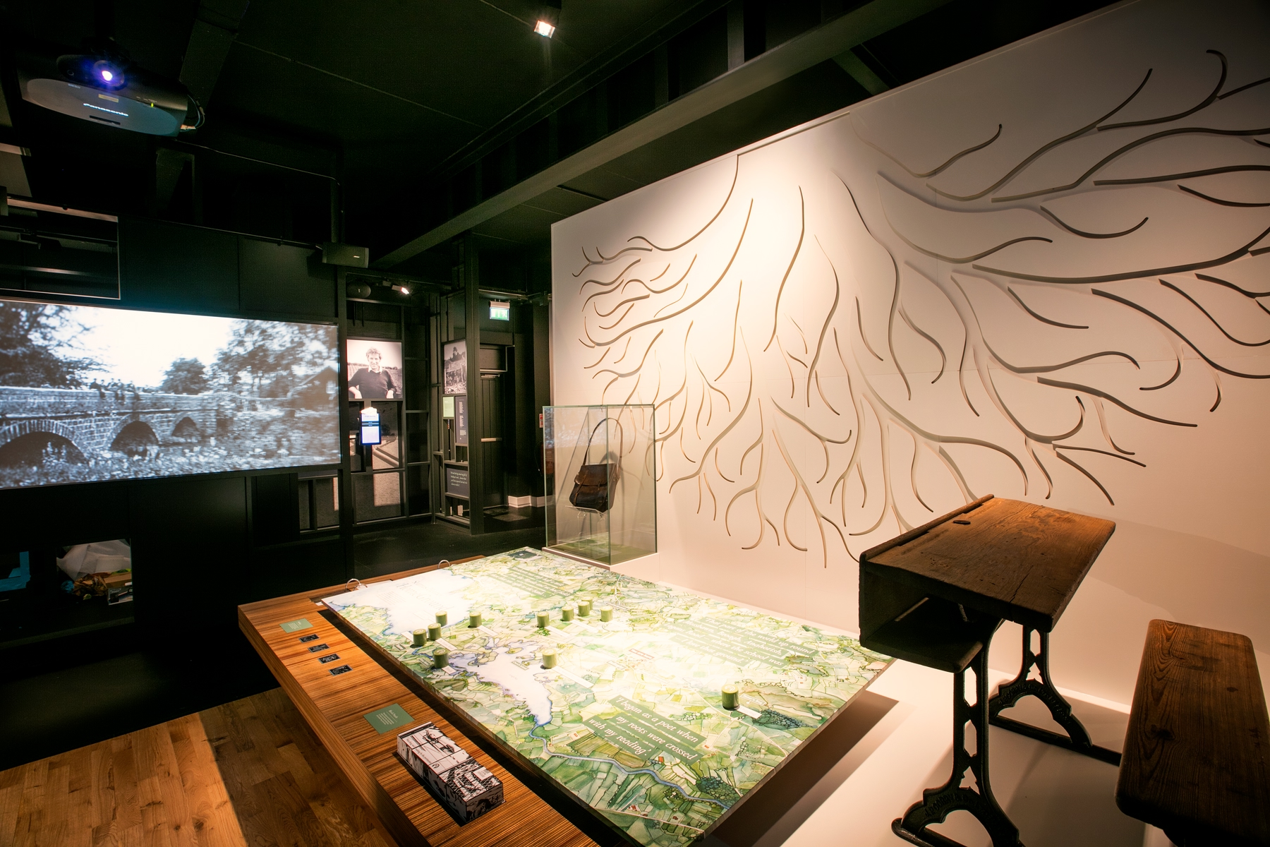 Interior of Seamus Heaney HomePlace, a museum dedicated to the famous Irish poet. The scene shows a wall painted with tree roots, an old-fashioned classroom desk and a map on a table.