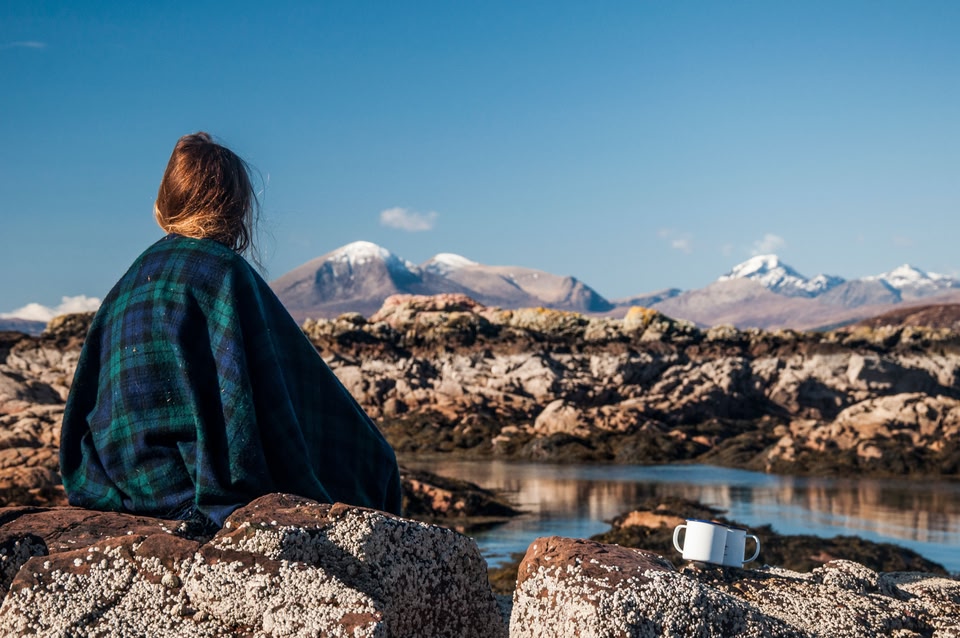 A woman sitting on a rock wrapped in a tartan blanket looking at the distant mountains of Applecross Peninsula, Lochaber, West Highlands.