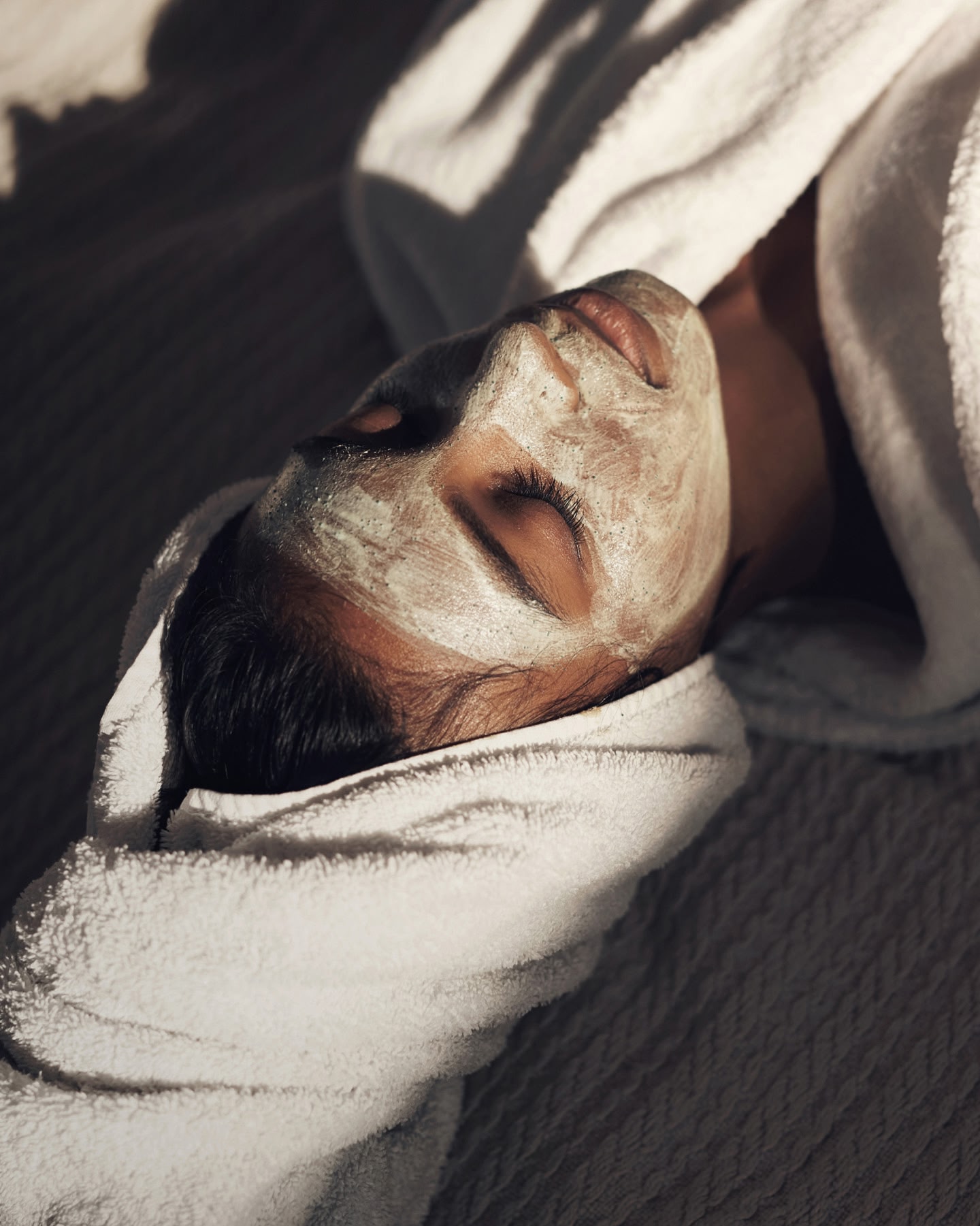  A woman wearing a white robe, lying down with a towel wrapped round her hair and a light green face mask on her face.