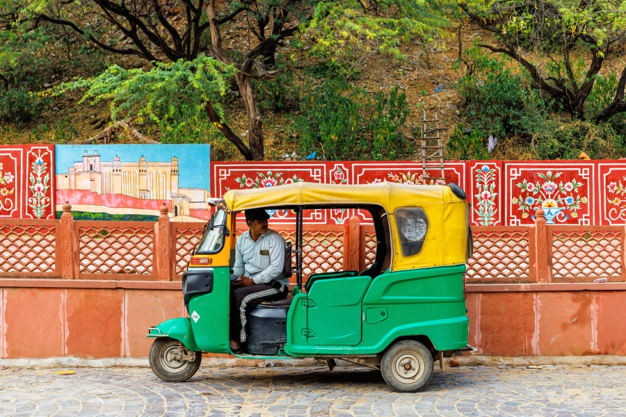 A green and yellow auto-rickshaw (tuk-tuk) driving along a street in Jaipur, with vibrantly painted red walls and a glimpse of a historic building visible in the background.
