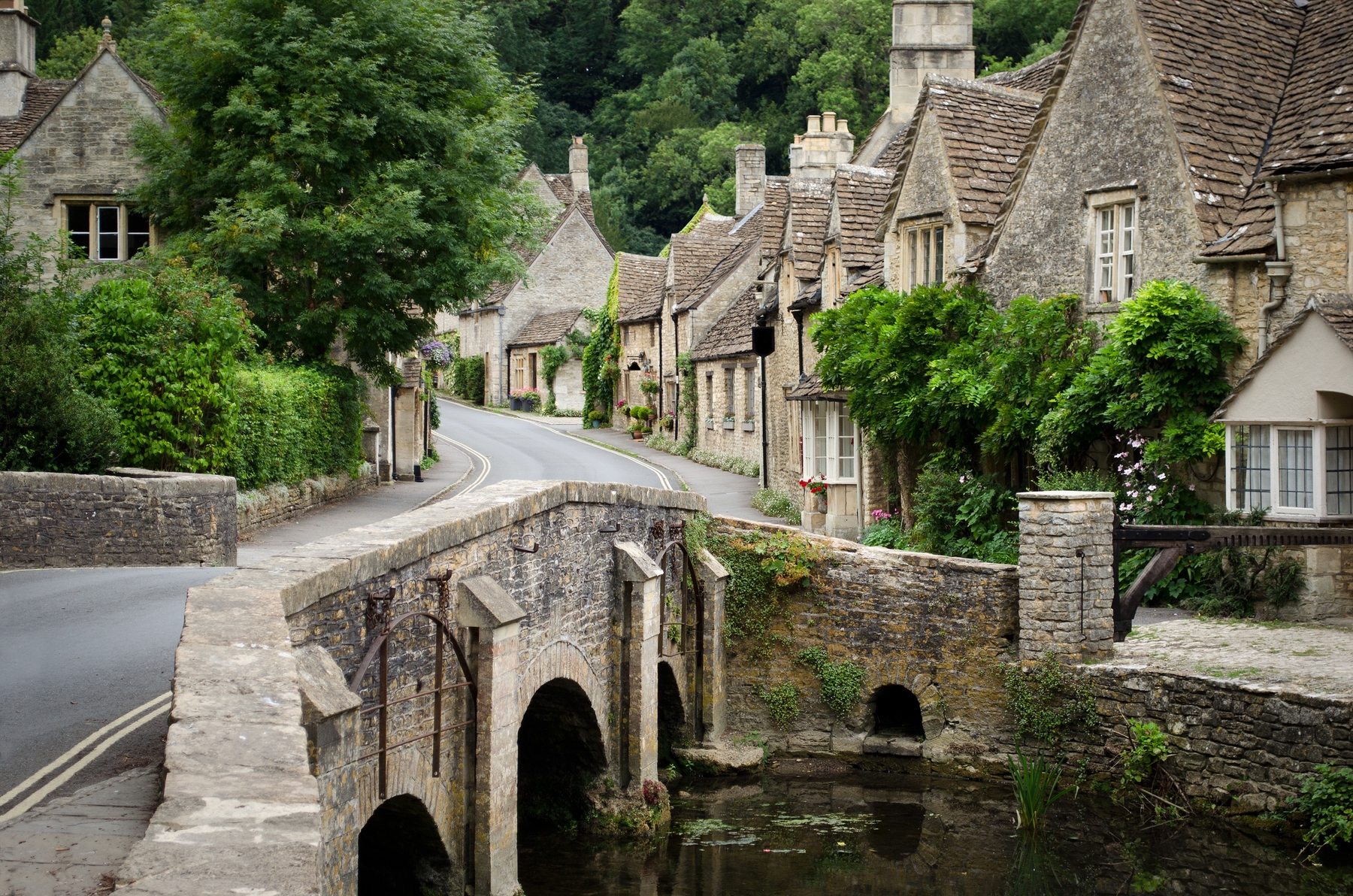 Pretty cottages line a street leading up to a stone bridge over a river in Castle Combe, a Cotswolds town perfect for spring holidays
