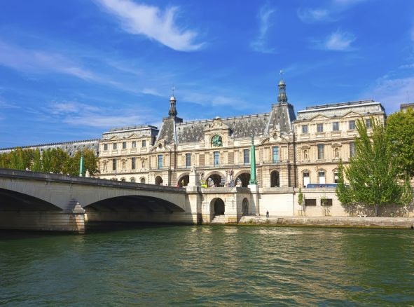 An image of an historical white and grey building near the river with a bridge.