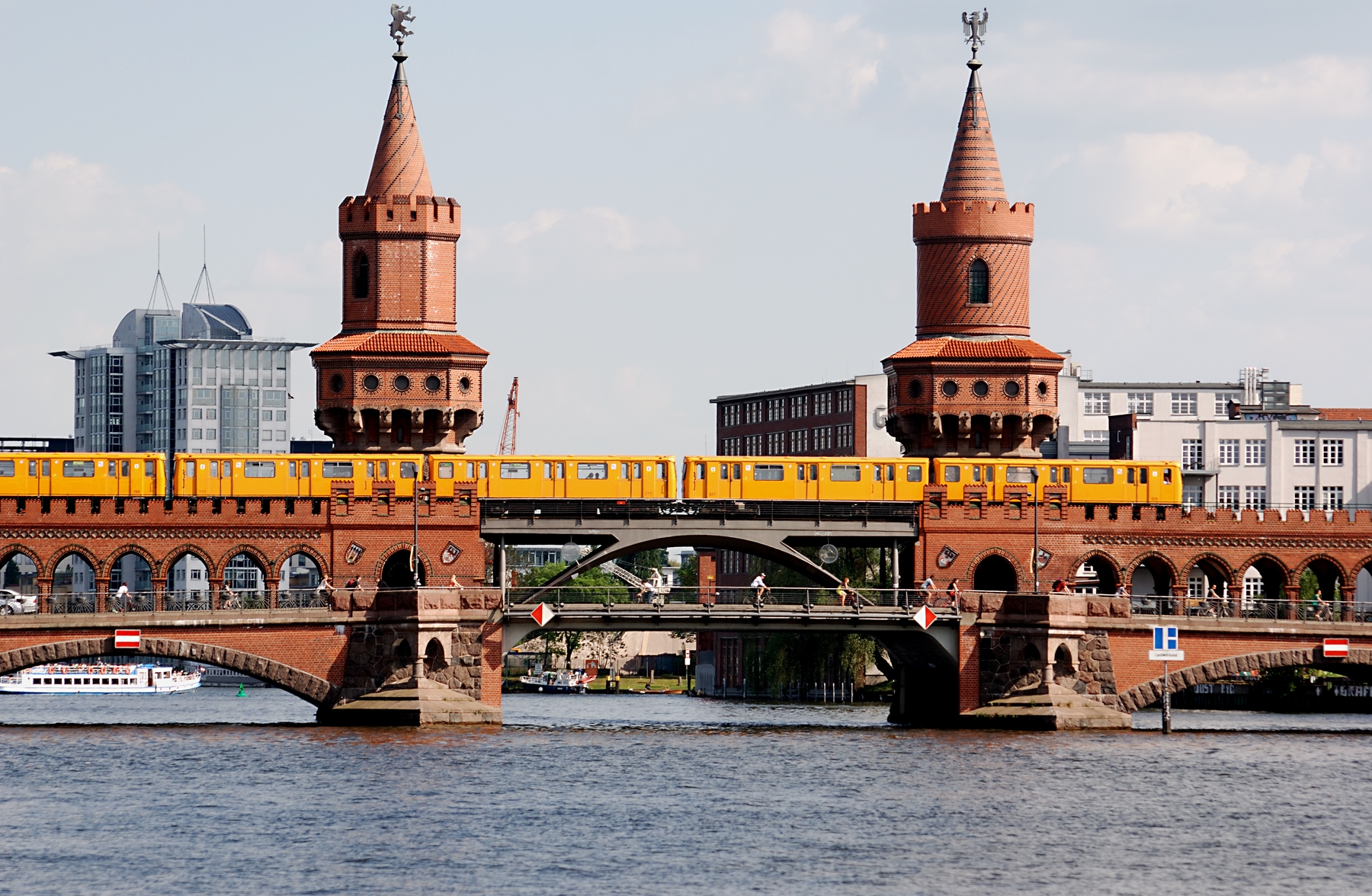 The double-decker Oberbaum Bridge in Berlin, crossing the River Spree. A yellow U-bahn train is gliding across the top level as people stroll underneath.
