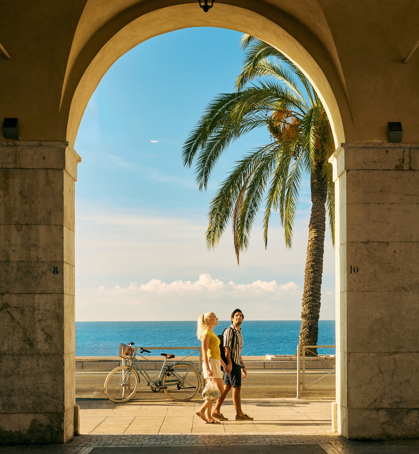 フランス、ニースの海辺で、ヤシの木々の横のアーチ道を散歩するカップル。