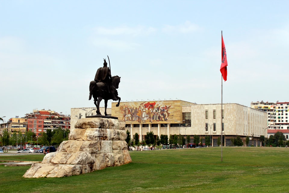 Statue of Albanian national hero Skanderbeg in Skanderbeg Square, Tirana, with the National History Museum and its socialist realist mural in the background, and the Albanian flag flying on a tall flagpole.