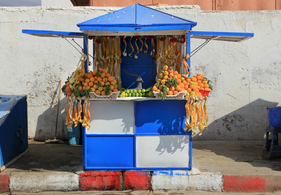 Colourful street fruit stall in Marrakech, decorated with hanging citrus peels and stacked with oranges, bananas, and limes, set against a whitewashed wall.