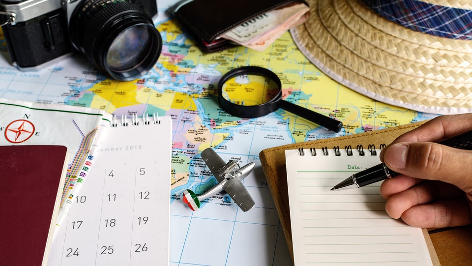 a travel planning scene. A world map serves as the backdrop, overlaid with various travel-related items such as a straw hat, a magnifying glass, a toy airplane, a calendar and a hand holding a pen.