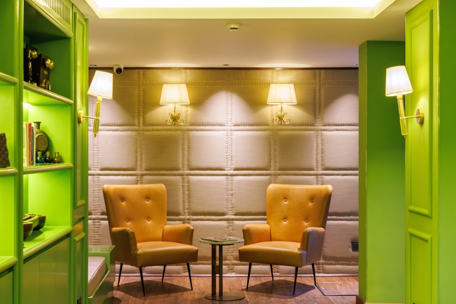 A cozy lounge nook at Abode Jaipur with two caramel-colored wingback armchairs flanking a small round table, set against a padded silver wall panel and framed by bright lime-green doorways and shelving.