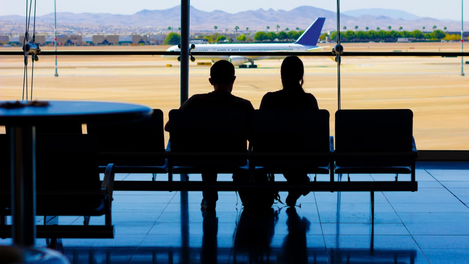 two people sat on airport chairs facing the window looking at a plane on the runway 