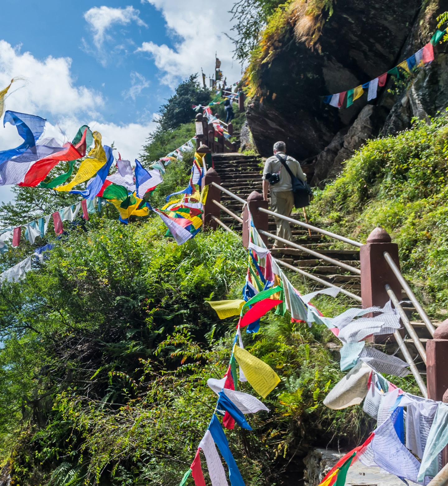 A man walks up stone stairs on the side of a mountain, bordered by green shrubbery and Tibetan prayer flags in Paro, Bhutan