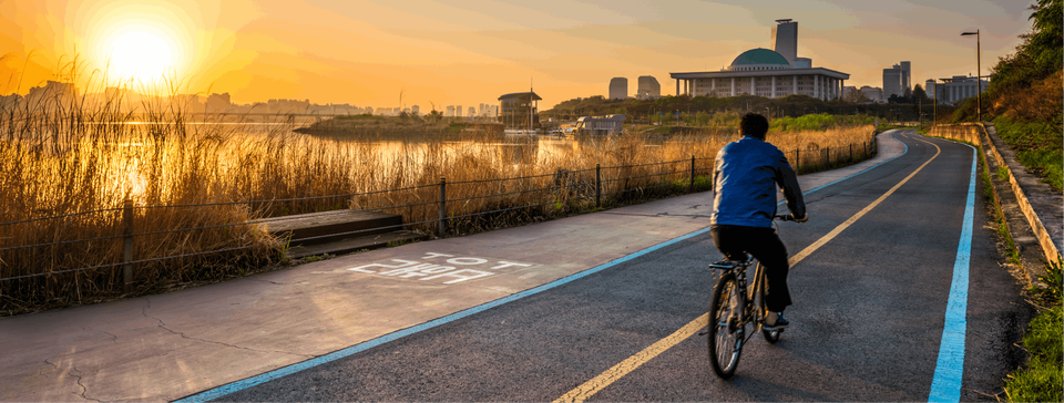 A man cycling along a scenic route, gazing to his left at a serene lake. In the distance, a golden sun sets, creating a picturesque scene.