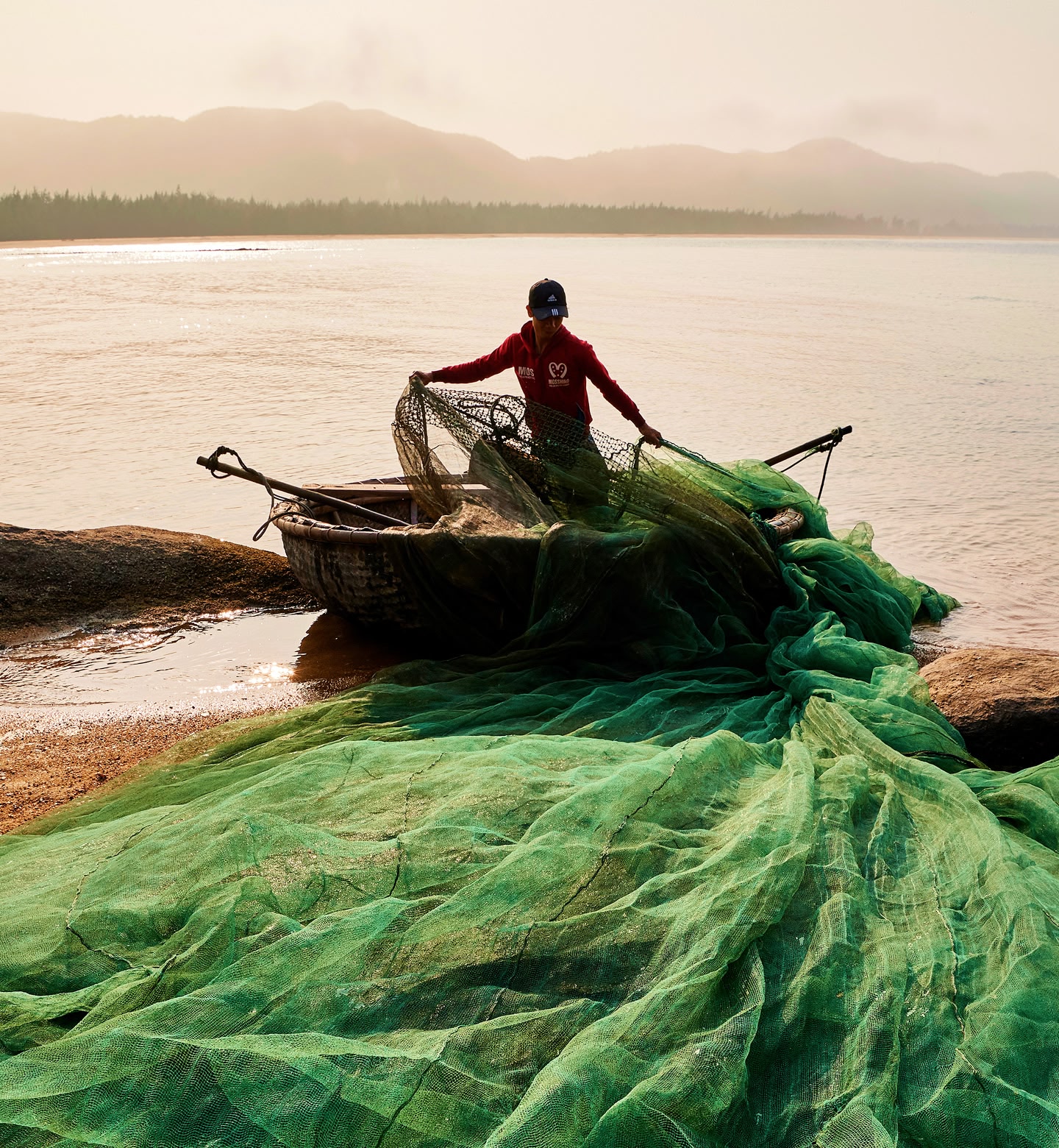 베트남 후에의 새벽 풍경. 둥근 나무 배 옆에서 초록색 그물을 펼치는 어부의 모습.